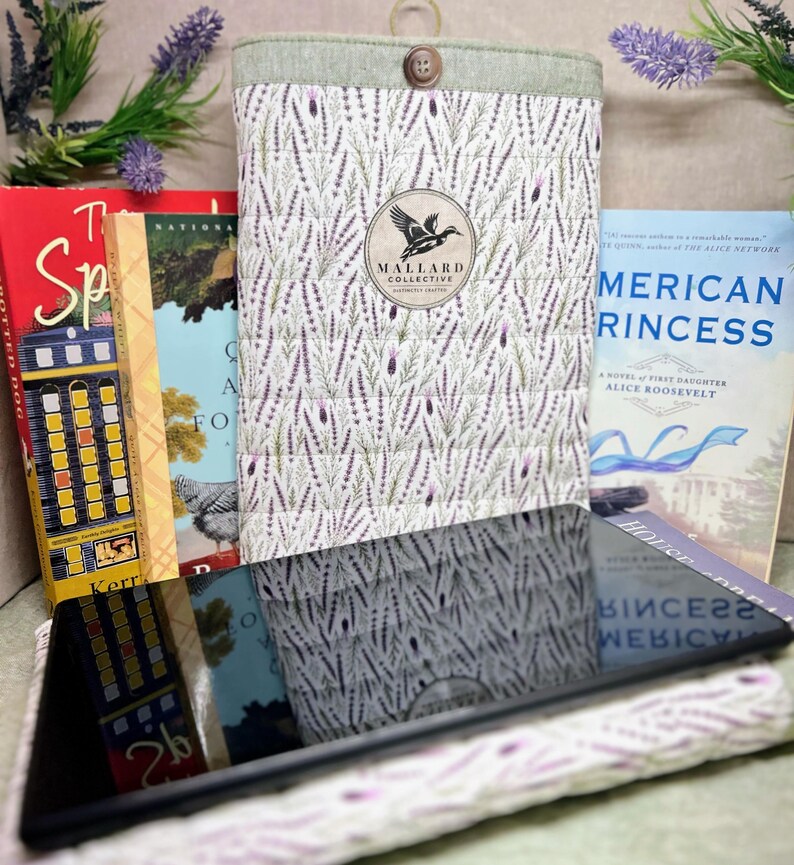 May include: A quilted fabric sleeve featuring a lavender sprig pattern, a light green top border with a single brown button, and a circular logo patch with a duck silhouette and the text Mallard Collective Distinctly Crafted.