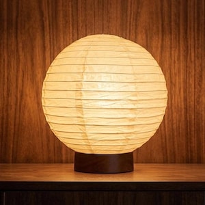 Op de afbeelding: Een ronde, crèmekleurige papieren lantaarnlamp met horizontale lijnen, rustend op een donkerbruine houten voet. De lamp is verlicht en geeft een warme gloed. De achtergrond is een donker houten paneel.
