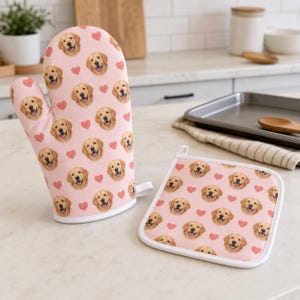 May include: A light pink oven mitt and a square pot holder featuring a repeating pattern of golden retriever dog faces and small red hearts. Both kitchen items have white trim and are made of fabric with a consistent printed design throughout.