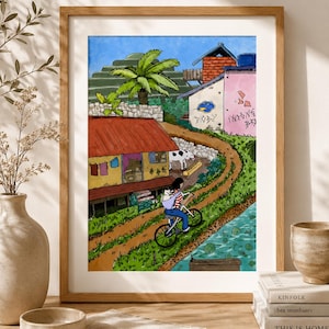 Pode incluir: Uma impressão artística emoldurada que retrata uma cena rural com uma pessoa a andar de bicicleta enquanto transporta um gato num suporte. A ilustração apresenta uma casa com telhado vermelho, um edifício rosa com graffiti, uma palmeira, uma vaca e um riacho.