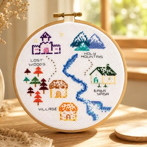 Pode incluir: Um bastidor de bordado em ponto cruz com um desenho de mapa. Apresenta um castelo, montanhas, árvores, casas e o texto "Lost Woods", "Holy Mountains", "Village" e "Baba Yaga". O desenho é bordado em tecido branco.