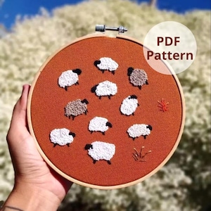 Pode incluir: Um bastidor de bordado com tecido laranja queimado com ovelhas bordadas. As ovelhas são brancas com rostos e pernas pretas, e algumas são marrons. O texto "PDF Pattern" está em um círculo branco.