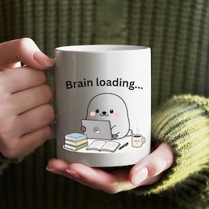 May include: White ceramic mug featuring the text "Brain loading..." and a cartoon seal using a laptop. The design includes books, a pen, and a coffee cup. The mug is held by a person wearing a green jumper.