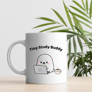 May include: White ceramic mug with the text "Tiny Study Buddy" and a cartoon seal using a laptop. A small coffee cup with the word "FOCUS" is next to the seal. The mug is on a light surface with green plant leaves in the background.