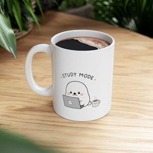 May include: White ceramic mug with a black coffee and a handle. The mug features a cartoon seal working on a laptop with the text "STUDY MODE" and a small coffee cup with the word "FOCUS". The mug is on a wooden surface.