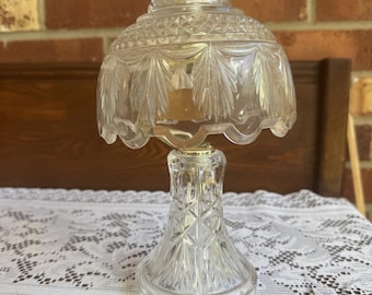 May include: A clear glass table lamp featuring a textured, diamond-patterned base and a matching scalloped glass shade with leaf-like carvings. The lamp is topped with a small, polished gold-tone metal finial and includes a white electrical cord.