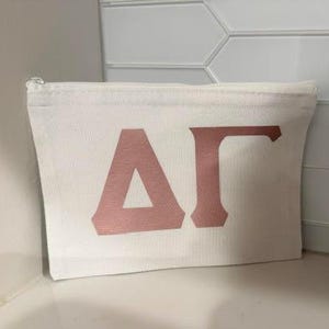 May include: A rectangular off-white canvas zipper pouch featuring the Greek letters Delta and Gamma in a metallic rose gold finish centered on the front. The pouch has a simple top zipper closure and a structured, flat-bottomed design.