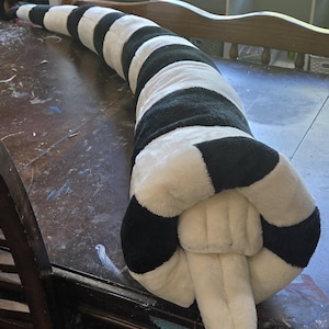 May include: A long, plush, stuffed tail accessory featuring alternating horizontal black and white stripes. The cylindrical, soft-textured item tapers toward the end and includes a smaller, white, pointed appendage extending from the base.