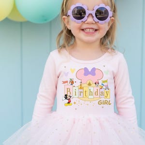 Può includere: Una bambina indossa una maglietta rosa chiaro a maniche lunghe e un tutù rosa. La maglietta ha una grafica con la scritta "Birthday Girl" con personaggi Disney. La bambina indossa anche occhiali da sole viola a forma di fiore.