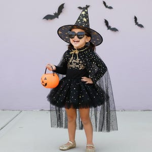 Könnte beinhalten: Ein Halloween-Hexenkostüm für Kinder. Das schwarze Outfit umfasst einen Tüllrock, ein langärmliges Oberteil mit goldenen Sternen und einen Umhang. Der Hexenhut hat goldene Sterne. Das Kind hält einen orangefarbenen Kürbis-Eimer und trägt herzförmige Sonnenbrillen. Der Text auf dem Shirt lautet "I put a spell on you."