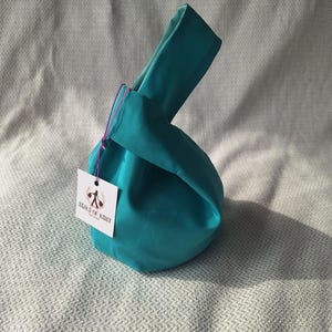 May include: A teal fabric knot bag with a single handle, featuring a smooth, solid-coloured exterior. A small white square tag is attached to the side with a thin purple string, displaying the text "SEALS OF KENT" and a graphic logo.