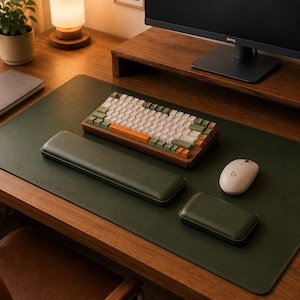 May include: A modern desk setup featuring a green desk mat, a mechanical keyboard with white, green, and orange keys, a white mouse, and a matching wrist rest. A wooden monitor stand and a notebook are also visible.