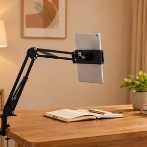 May include: A black tablet holder with an adjustable arm is clamped to a wooden desk. A silver tablet is secured in the holder. A notebook and pen are on the desk. A lamp and a potted plant are in the background.