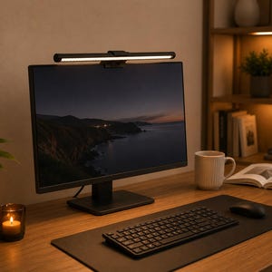 Può includere: Un monitor di computer nero con una lampada da scrivania attaccata sopra. Una tastiera, un mouse e un tappetino nero sono davanti al monitor su una scrivania di legno. Una candela accesa, una tazza e un libro aperto sono anche sulla scrivania.