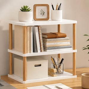 May include: A three-tier desktop organizer featuring white shelves supported by light wood-toned cylindrical pillars. The unit holds various books, a white storage box, a small potted succulent, a wooden picture frame, and two pen holders.
