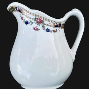 May include: White ceramic creamer with a floral design in shades of pink, blue, and gold. The creamer has a scalloped rim and a handle.