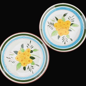 May include: Two white ceramic plates with a floral design. Each plate features a yellow flower bouquet in the center, surrounded by green leaves and black stems. The rims have a blue and green border.