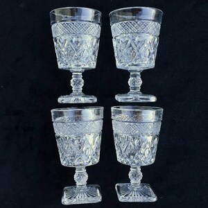 May include: Four clear, vintage water goblets with intricate diamond and geometric patterns. The glasses have a decorative band near the rim and sit on a pedestal base. The bases are square-shaped.