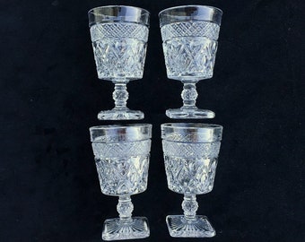May include: Four clear, vintage water goblets with intricate diamond and geometric patterns. The glasses have a decorative band near the rim and sit on a pedestal base. The bases are square-shaped.