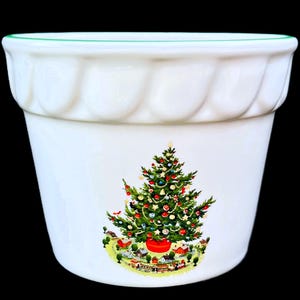 May include: White ceramic planter with a scalloped rim and a green accent. The planter features a colorful Christmas tree design with ornaments and a village scene at the base. A festive holiday decoration.