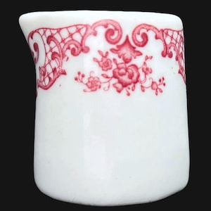 Puede incluir: Una pequeña jarra de cerámica blanca con un diseño floral y enrejado rojo alrededor del borde. La jarra tiene un asa curva y un pico para verter fácilmente. El diseño presenta flores y adornos decorativos.