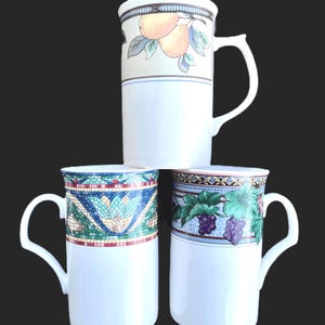 May include: Three white ceramic mugs with decorative bands. One mug features peaches, another a mosaic pattern, and the third has grapes. Each mug has a white handle and a black background.