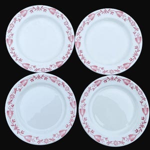 May include: Four white ceramic plates with a red floral border design. The plates have a simple, elegant style, with a repeating pattern of stylized flowers and leaves around the rim. The plates are arranged on a black background.