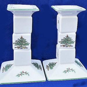 May include: A pair of white ceramic candle holders with a green trim and a Christmas tree design. The candle holders are stacked in a square shape.