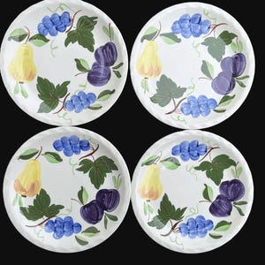 May include: Four white ceramic plates with a hand-painted fruit design. Each plate features a yellow pear, blue grapes, and purple plums, surrounded by green leaves and vines. The plates have a textured, wavy rim.