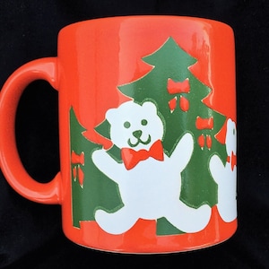 May include: Orange ceramic mug with a Christmas theme. The mug features two white teddy bears with red bows, standing in front of green Christmas trees with red bows and ornaments.