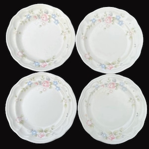 May include: Four white ceramic plates with scalloped edges. Each plate features a floral design with pink roses, blue flowers, and green leaves. The plates are arranged on a black background.
