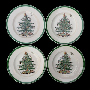 May include: Four white ceramic plates with a green border. Each plate features a detailed illustration of a Christmas tree decorated with ornaments and gifts. The plates are arranged on a black background.