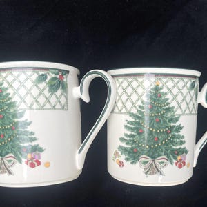 May include: Two off-white ceramic mugs featuring a green Christmas tree design with red ornaments, a gold star topper, and a bow at the base. The upper rim displays a green lattice pattern with holly leaves. Each mug has a curved handle with green trim.