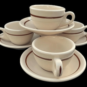 May include: A set of four cream-colored ceramic cups and matching saucers, each featuring a single horizontal brown stripe around the center of the cup and a matching brown circular band on the rim of the saucer, arranged in a cluster.