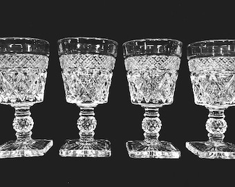 May include: Four clear crystal wine glasses with diamond and geometric patterns. Each glass has a square base, a beaded stem, and a detailed bowl. The glasses are arranged in a row against a dark background, showcasing their intricate design.