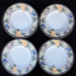 May include: Four white ceramic plates with a fruit design. Each plate features a border with illustrations of pears, grapes, and leaves in shades of yellow, blue, and purple. The plates have a light green background and a dark brown rim.