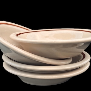 May include: A stack of three off-white ceramic bowls, each featuring a single thin brown band encircling the inner rim. The bowls are arranged with one tilted at an angle, showcasing the smooth, glossy finish and the simple, rounded design.
