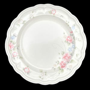 May include: Off-white ceramic plate with a scalloped edge. The plate is decorated with pink and blue floral patterns and green leaves. The floral design is arranged around the rim of the plate.