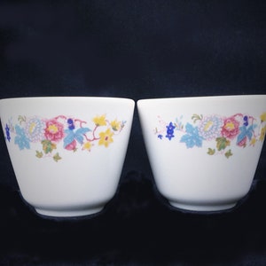 May include: Two white ceramic bowls with a floral design. The design features pink, blue, yellow, and green flowers and leaves. The bowls have a simple, elegant shape and are set against a dark background.