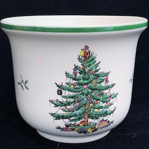 May include: A white ceramic Christmas planter with a green rim. The planter features a detailed illustration of a decorated Christmas tree with Santa Claus on top, surrounded by wrapped gifts. Small sprigs of mistletoe are also visible.