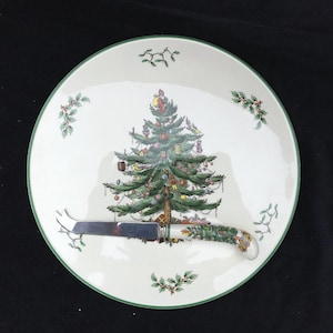 May include: A white Christmas-themed plate with a green rim and a decorated Christmas tree in the center. A cheese knife with a festive handle rests on the plate. Holly and mistletoe accents are scattered around the plate.