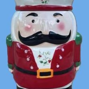 May include: A ceramic nutcracker cookie jar featuring a red jacket with white buttons, a black belt with a yellow buckle, and a green hat. The figure has a black mustache, rosy cheeks, and a sprig of holly painted on the chest and hat brim.