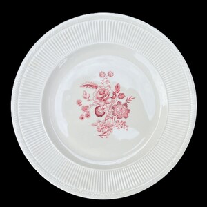 May include: A round white ceramic dinner plate featuring a central floral motif in a deep red color. The plate has a wide, raised rim decorated with a series of vertical, parallel ridges that extend around the entire circumference of the edge.