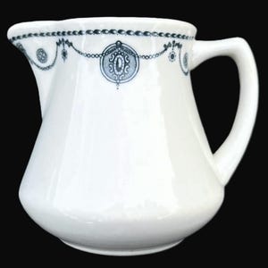 May include: White ceramic pitcher with a curved handle and a decorative blue and white design around the rim. The design includes a chain of circles and oval medallions. The pitcher has a rounded body and a pouring spout.