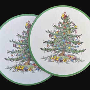 May include: Two white ceramic Christmas tree plates with green trim. Each plate features a green Christmas tree with colorful ornaments, presents, and a small figure at the top.