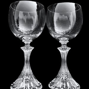 May include: Two clear crystal wine glasses featuring large, rounded bowls and intricate, vertically fluted stems that flare out into wide, faceted bases. The glass surfaces are smooth and transparent, reflecting light against a black background.