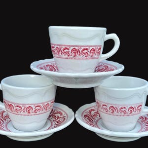 May include: Three white ceramic teacups and saucers with a red floral pattern. The cups and saucers have a scalloped edge.