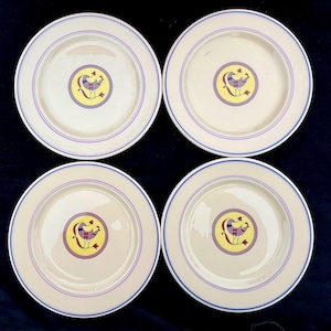 May include: Four cream-colored plates with a blue and purple border. Each plate features a central design of a bird in yellow, purple, and red. The plates are arranged on a dark blue surface.