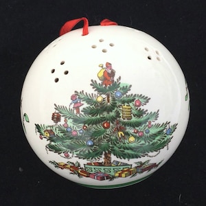 May include: A white ceramic Christmas ornament shaped like a sphere with a red ribbon. The ornament features a green Christmas tree with colorful ornaments, presents, and a small figure of Santa Claus.