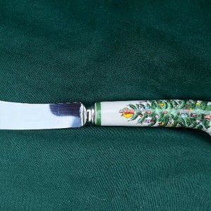May include: A butter knife with a stainless steel blade and a white handle decorated with a Christmas tree design. The handle has a green band near the blade and a curved end.